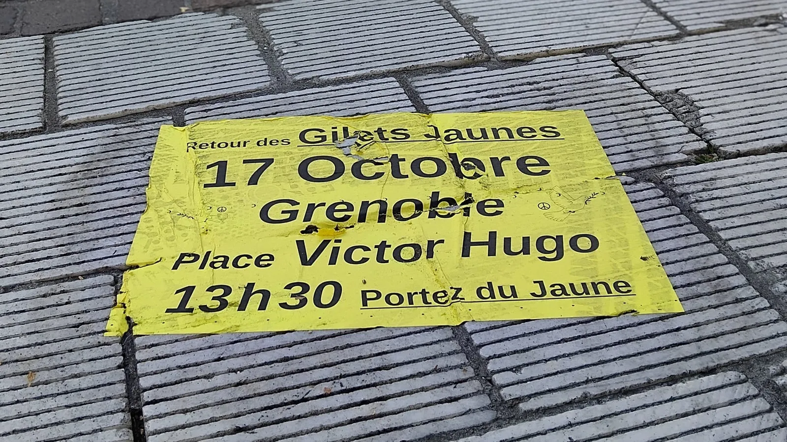 Volantino giallo a terra su un marciapiede di Grenoble: Retour des Gilets Jaunes, 17 ottobre, place Victor Hugo, ore 13.30